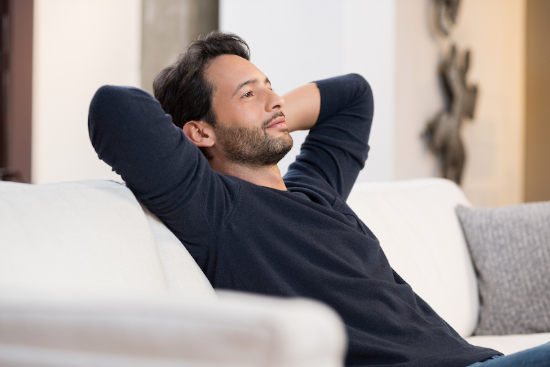 Portrait of a young man resting on sofa and thinking about the future. Handsome young man with hands behind head sitting on couch in living room. Positive man daydreaming and relaxing at home.